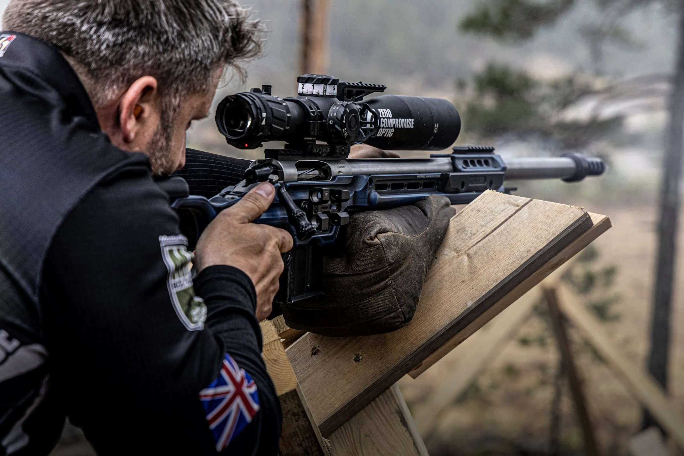 Josh Martin behind a Zero Compromise Optic scope, Union Jack on his sleeve, muzzle blast in the air